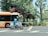 A motorcyclist wearing a white helmet and blue uniform rides a police motorcycle in front of an orange and cream city bus. The scene is set in an urban area with lush green trees and foliage in the background. A black car is partially visible behind the bus. The pavement is marked with white road crossing lines.