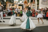 Four performers wearing colorful and ornate costumes engage in a lively dance in front of a crowd. The crowd, consisting of adults and children, watches attentively, some holding up phones to capture the moment. Decorative, illuminated arches and structures are visible in the background, suggesting a festive or celebratory event.