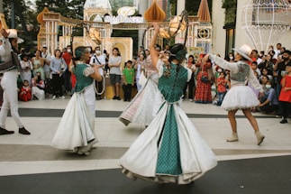 Four performers wearing colorful and ornate costumes engage in a lively dance in front of a crowd. The crowd, consisting of adults and children, watches attentively, some holding up phones to capture the moment. Decorative, illuminated arches and structures are visible in the background, suggesting a festive or celebratory event.