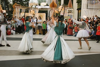 Four performers wearing colorful and ornate costumes engage in a lively dance in front of a crowd. The crowd, consisting of adults and children, watches attentively, some holding up phones to capture the moment. Decorative, illuminated arches and structures are visible in the background, suggesting a festive or celebratory event.