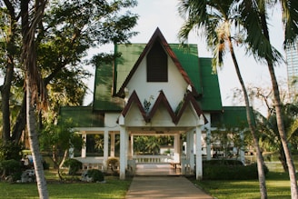 A building with a triangular roof, featuring green and red accents, surrounded by lush greenery and palm trees. There is a pathway leading to the entrance, and the area seems calm and serene.