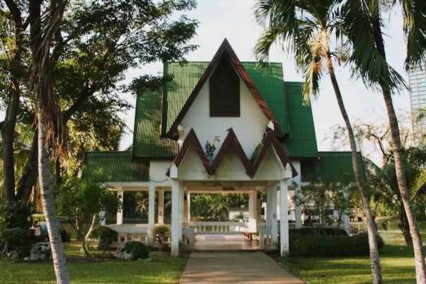 A building with a triangular roof, featuring green and red accents, surrounded by lush greenery and palm trees. There is a pathway leading to the entrance, and the area seems calm and serene.
