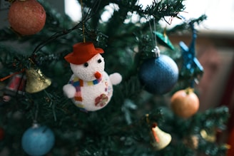 A Christmas tree decorated with various colorful ornaments, including a snowman figure wearing a red hat and a scarf, hanging alongside blue, orange, and gold baubles. The tree is lush and green, suggesting a festive holiday atmosphere.