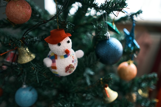 A Christmas tree decorated with various colorful ornaments, including a snowman figure wearing a red hat and a scarf, hanging alongside blue, orange, and gold baubles. The tree is lush and green, suggesting a festive holiday atmosphere.
