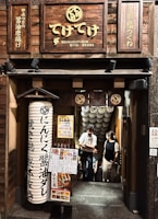 Entrance to a Japanese restaurant with wooden signage featuring Japanese characters. A large paper lantern with more Japanese writing is positioned outside the entrance. Several people are seen descending a staircase behind the entrance, with menus and posters advertising food and drink specials displayed prominently.