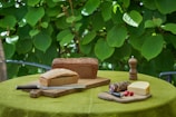 A rustic outdoor setting features a table with a green tablecloth, adorned with loaves of freshly baked bread on wooden boards. Slices of cheese and cured sausage are placed nearby, accompanied by a knife, two small radishes, and a wooden pepper mill. The background is lush with large green leaves.