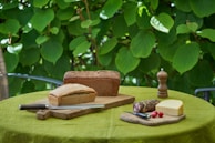A rustic outdoor setting features a table with a green tablecloth, adorned with loaves of freshly baked bread on wooden boards. Slices of cheese and cured sausage are placed nearby, accompanied by a knife, two small radishes, and a wooden pepper mill. The background is lush with large green leaves.