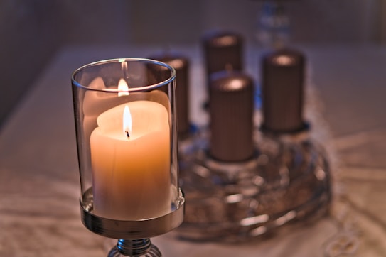 A large candle encased in a glass holder is prominently displayed in the foreground, with a soft, warm flame. In the background, several unlit pillar candles are placed on a decorative metallic tray, arranged in a circular formation. The scene is set on a light-colored tablecloth, creating a calm and serene atmosphere.