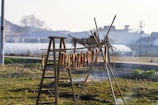 Open field with strips of meat hanging on a wooden drying rack. There's a wooden ladder positioned next to the rack. The background includes greenhouses, houses, and a faint mist in the air, suggesting early morning or late afternoon light.