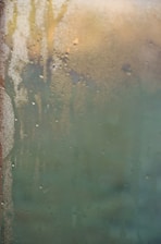 Condensation covers a glass surface with water droplets, creating a blurred effect. In the background, vague shapes and colors suggest a possible scene or objects obscured by the condensation.