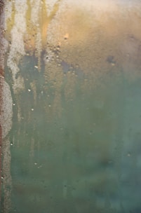 Condensation covers a glass surface with water droplets, creating a blurred effect. In the background, vague shapes and colors suggest a possible scene or objects obscured by the condensation.