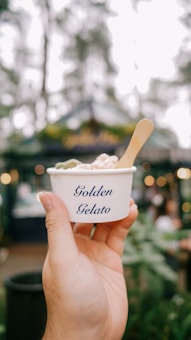 A hand is holding a small white cup labeled 'Golden Gelato' with a spoon, in an outdoor setting. The background is blurred, showing greenery and soft lights, suggesting a casual and relaxed environment.