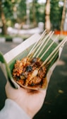 A hand holds a serving of grilled skewered meat, likely satay, wrapped in a green banana leaf. The skewers appear charred and covered with a spicy, brown sauce. The background is a blurred outdoor setting with greenery.