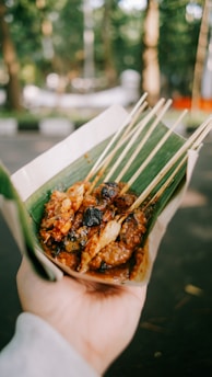 A hand holds a serving of grilled skewered meat, likely satay, wrapped in a green banana leaf. The skewers appear charred and covered with a spicy, brown sauce. The background is a blurred outdoor setting with greenery.