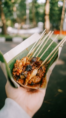 A hand holds a serving of grilled skewered meat, likely satay, wrapped in a green banana leaf. The skewers appear charred and covered with a spicy, brown sauce. The background is a blurred outdoor setting with greenery.