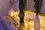 A decorative setup on a dresser features two white vases with tall green plants and a tall dark glass vase. In the center, two illuminated gift-wrapped light decorations create a warm glow. A gold-framed mirror reflects the scene, enhancing the ambient light. Delicate ornamental branches with small spherical elements are spread across the surface, adding a festive touch.