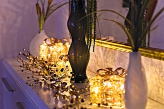 A decorative setup on a dresser features two white vases with tall green plants and a tall dark glass vase. In the center, two illuminated gift-wrapped light decorations create a warm glow. A gold-framed mirror reflects the scene, enhancing the ambient light. Delicate ornamental branches with small spherical elements are spread across the surface, adding a festive touch.
