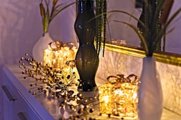 A decorative setup on a dresser features two white vases with tall green plants and a tall dark glass vase. In the center, two illuminated gift-wrapped light decorations create a warm glow. A gold-framed mirror reflects the scene, enhancing the ambient light. Delicate ornamental branches with small spherical elements are spread across the surface, adding a festive touch.
