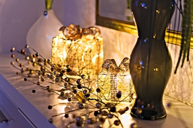 Two decorative, illuminated gift boxes covered with golden beads are placed on a white surface. Beside them, a glossy, dark-colored vase holds long branches. A vine-like string with small golden and dark beads is artistically arranged around the gifts. The soft, warm glow from the lights creates a cozy and elegant atmosphere.