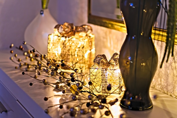 Two decorative, illuminated gift boxes covered with golden beads are placed on a white surface. Beside them, a glossy, dark-colored vase holds long branches. A vine-like string with small golden and dark beads is artistically arranged around the gifts. The soft, warm glow from the lights creates a cozy and elegant atmosphere.