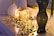 Two decorative, illuminated gift boxes covered with golden beads are placed on a white surface. Beside them, a glossy, dark-colored vase holds long branches. A vine-like string with small golden and dark beads is artistically arranged around the gifts. The soft, warm glow from the lights creates a cozy and elegant atmosphere.