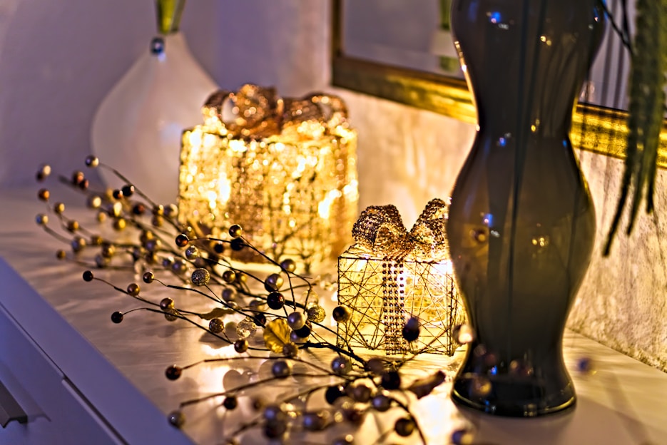 Two decorative, illuminated gift boxes covered with golden beads are placed on a white surface. Beside them, a glossy, dark-colored vase holds long branches. A vine-like string with small golden and dark beads is artistically arranged around the gifts. The soft, warm glow from the lights creates a cozy and elegant atmosphere.