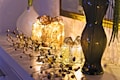 Two decorative, illuminated gift boxes covered with golden beads are placed on a white surface. Beside them, a glossy, dark-colored vase holds long branches. A vine-like string with small golden and dark beads is artistically arranged around the gifts. The soft, warm glow from the lights creates a cozy and elegant atmosphere.