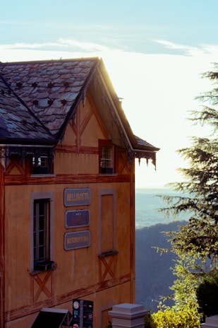A charming building with rustic architecture, featuring a weathered roof and ornate wooden details. The signs on the exterior read 'Bellavista' and 'Ristorante Bar'. The structure is set against a backdrop of a scenic landscape with trees and a distant horizon under a bright sky.