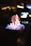 A close-up of a rectangular dish with a flaming dessert, possibly a brûlée or flambé. The flames are predominantly blue with hints of orange and yellow. The background is blurred, with bokeh light effects in warm tones, suggesting a dimly lit or nighttime setting.