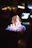 A close-up of a rectangular dish with a flaming dessert, possibly a brûlée or flambé. The flames are predominantly blue with hints of orange and yellow. The background is blurred, with bokeh light effects in warm tones, suggesting a dimly lit or nighttime setting.