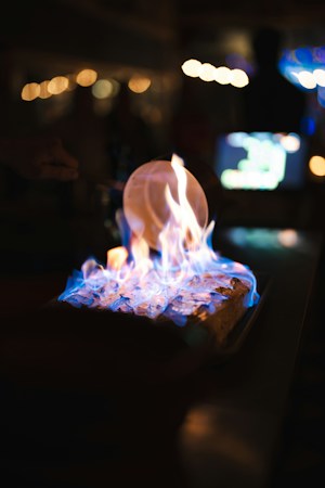 A close-up of a rectangular dish with a flaming dessert, possibly a br&ucirc;l&eacute;e or flamb&eacute;. The flames are predominantly blue with hints of orange and yellow. The background is blurred, with bokeh light effects in warm tones, suggesting a dimly lit or nighttime setting.