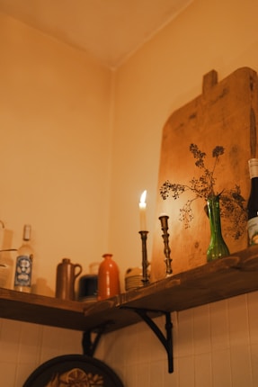 A warmly lit corner of a room showing a wooden shelf mounted on a wall, displaying a variety of objects including bottles, a candle on a brass holder, a green vase with dried flowers, and a wooden cutting board. The ambiance suggests a cozy, rustic setting.