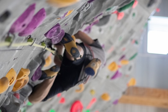 A person is climbing a rock wall indoors, using colorful climbing holds. The wall is vertical with holds of various shapes and colors, including purple, yellow, and green. The climber appears to be wearing specialized climbing shoes and is focused on ascent.