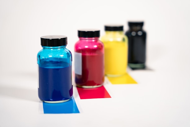 A set of four glass jars filled with colorful liquids is displayed. Each jar has a black cap and they contain distinct colors: blue, red, yellow, and black. The jars are aligned diagonally on a white surface, each placed on a colored strip matching the liquid inside.