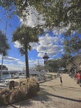 A marina with a few yachts docked along the water is visible. In the background, a red and white striped lighthouse stands tall against a bright blue sky with scattered clouds. Trees with green foliage frame the image, casting shadows on the brick pathway where people, including a child, are walking and sitting.
