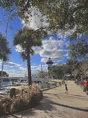 A marina with a few yachts docked along the water is visible. In the background, a red and white striped lighthouse stands tall against a bright blue sky with scattered clouds. Trees with green foliage frame the image, casting shadows on the brick pathway where people, including a child, are walking and sitting.