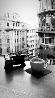 A black and white photograph captures a serene urban scene viewed from a high vantage point. A camera and a cup of coffee sit on a table next to a window, overlooking a street lined with classic and modern buildings. The outdoor scene includes people walking on the sidewalk below.