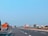 An empty highway stretches into the distance under a clear blue sky, with multiple colorful trucks parked along the side. Sparse greenery and utility poles line the road, giving the scene a sense of stillness.