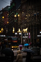 A street scene featuring a city bus with a display reading 'C Classic Line Laufener Kirchweg'. Pedestrians carrying umbrellas walk through a rainy setting, surrounded by buildings adorned with holiday lights and decorations depicting stars and festive figures.