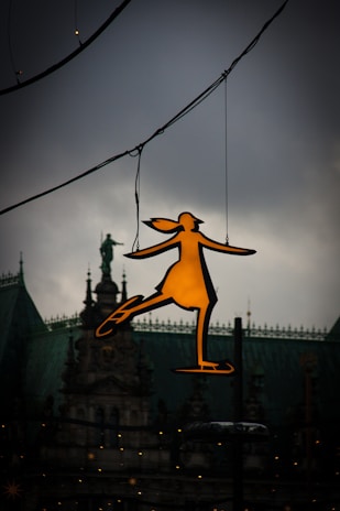 A silhouette of a girl on ice skates is illuminated and suspended in the foreground, set against a backdrop of a historic or ornate building with architectural details. The sky is overcast, creating a contrast between the bright figure and the moody backdrop.