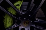 A close-up of a black alloy wheel featuring a lime green brake caliper with the word 'Porsche' written on it. The central hubcap displays a colorful crest insignia.