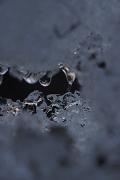 A close-up view of melting ice with intricate patterns and textures. The ice appears partially translucent, allowing light to reflect and refract through it, creating a dynamic visual effect. Several droplets hang from the edges, adding to the impression of thawing.
