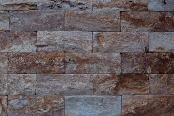 The image features a close-up view of a stone wall with a rectangular brick pattern. The bricks vary in shades of grey and brown, showcasing a rough, textured surface with visible grain and natural imperfections.