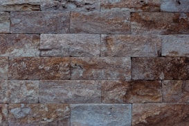 The image features a close-up view of a stone wall with a rectangular brick pattern. The bricks vary in shades of grey and brown, showcasing a rough, textured surface with visible grain and natural imperfections.