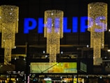 A large sign displaying the word 'PHILIPS' in bright blue letters atop a building. Below are decorative, vertically hanging lights that form cylinders made up of numerous tiny golden lights, creating a festive appearance. The background is a dimly lit urban setting with several small advertisements and signs visible, suggesting a commercial environment.
