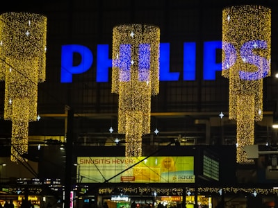 A large sign displaying the word 'PHILIPS' in bright blue letters atop a building. Below are decorative, vertically hanging lights that form cylinders made up of numerous tiny golden lights, creating a festive appearance. The background is a dimly lit urban setting with several small advertisements and signs visible, suggesting a commercial environment.