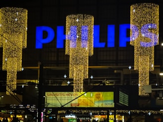 A large sign displaying the word 'PHILIPS' in bright blue letters atop a building. Below are decorative, vertically hanging lights that form cylinders made up of numerous tiny golden lights, creating a festive appearance. The background is a dimly lit urban setting with several small advertisements and signs visible, suggesting a commercial environment.
