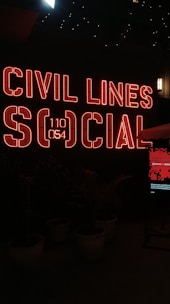 A vibrant neon sign with the words 'Civil Lines Social' is illuminated in bold red letters against a dark background. Below the sign, there are large ornate plant pots with greenery, and a digital screen displaying a message. The ambiance is modern and urban with a nightlife vibe.