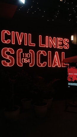 A vibrant neon sign with the words 'Civil Lines Social' is illuminated in bold red letters against a dark background. Below the sign, there are large ornate plant pots with greenery, and a digital screen displaying a message. The ambiance is modern and urban with a nightlife vibe.