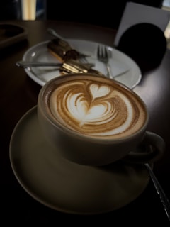 A cup of coffee with intricate latte art sits in the foreground on a saucer, featuring a heart and leaf design in the foam. In the background, a partially visible dessert, possibly a cake, is placed on a white plate with a fork and knife. The lighting is dim, creating a cozy and warm atmosphere.